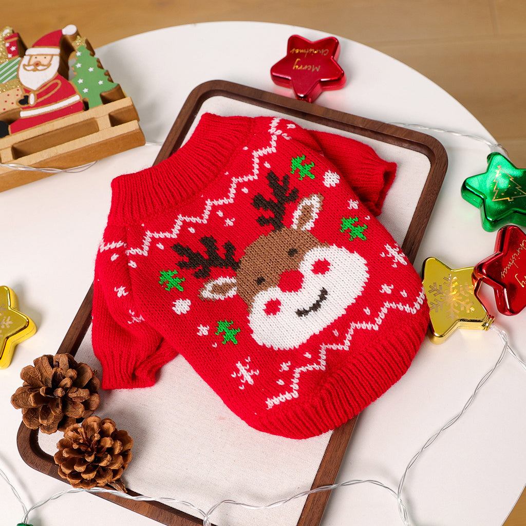 Red knitted dog sweater with reindeer design on a white surface with Christmas decorations.
