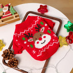 Red knitted dog sweater with reindeer design on a white surface with Christmas decorations.