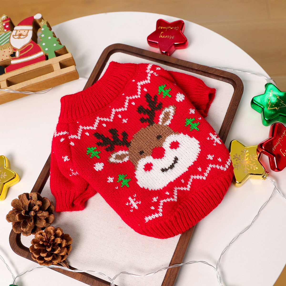 Red knitted dog sweater with reindeer design on a white surface with Christmas decorations.