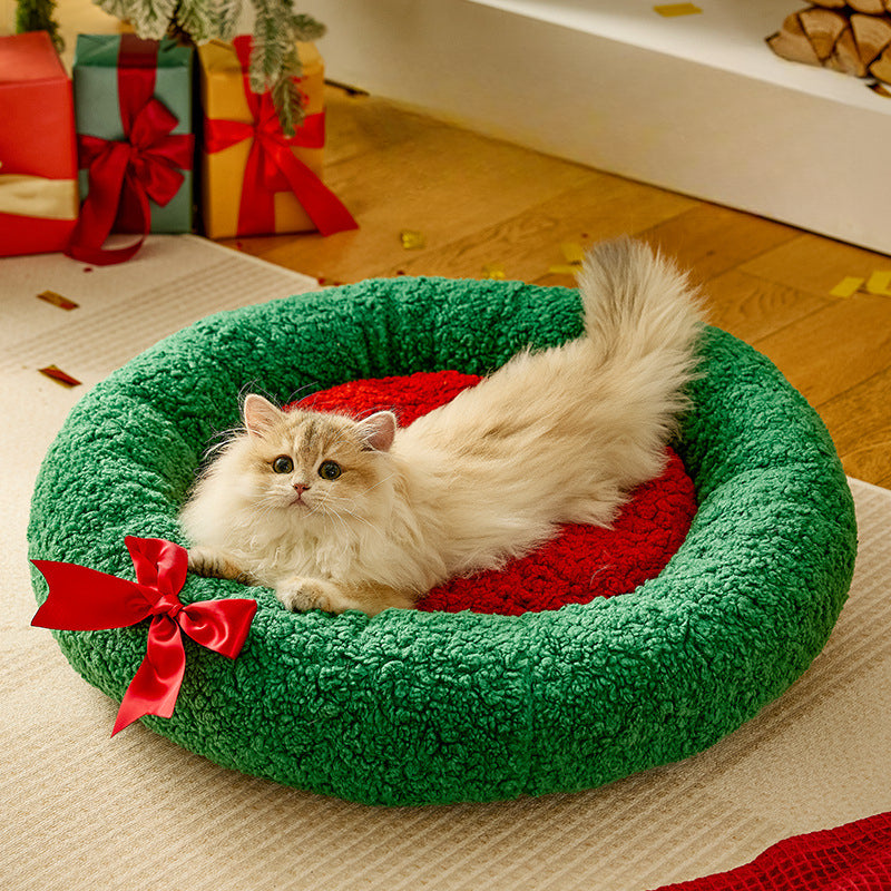 Cat lying on a green wreath-shaped pet bed with a red bow, surrounded by Christmas presents.