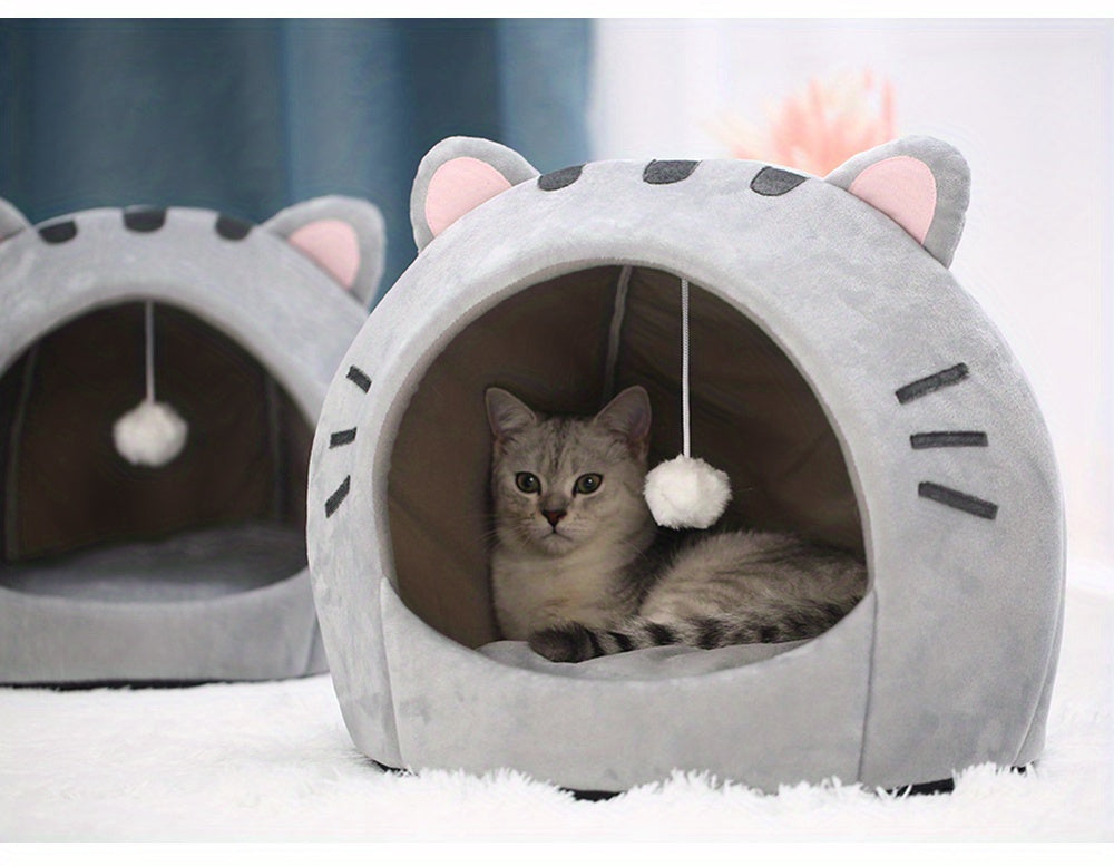 Cat lying inside a gray cat-shaped pet bed with a fluffy ball hanging inside.