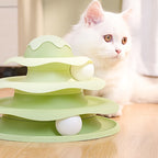 Green three-tiered cat toy with a white cat sitting next to it on a wooden surface.