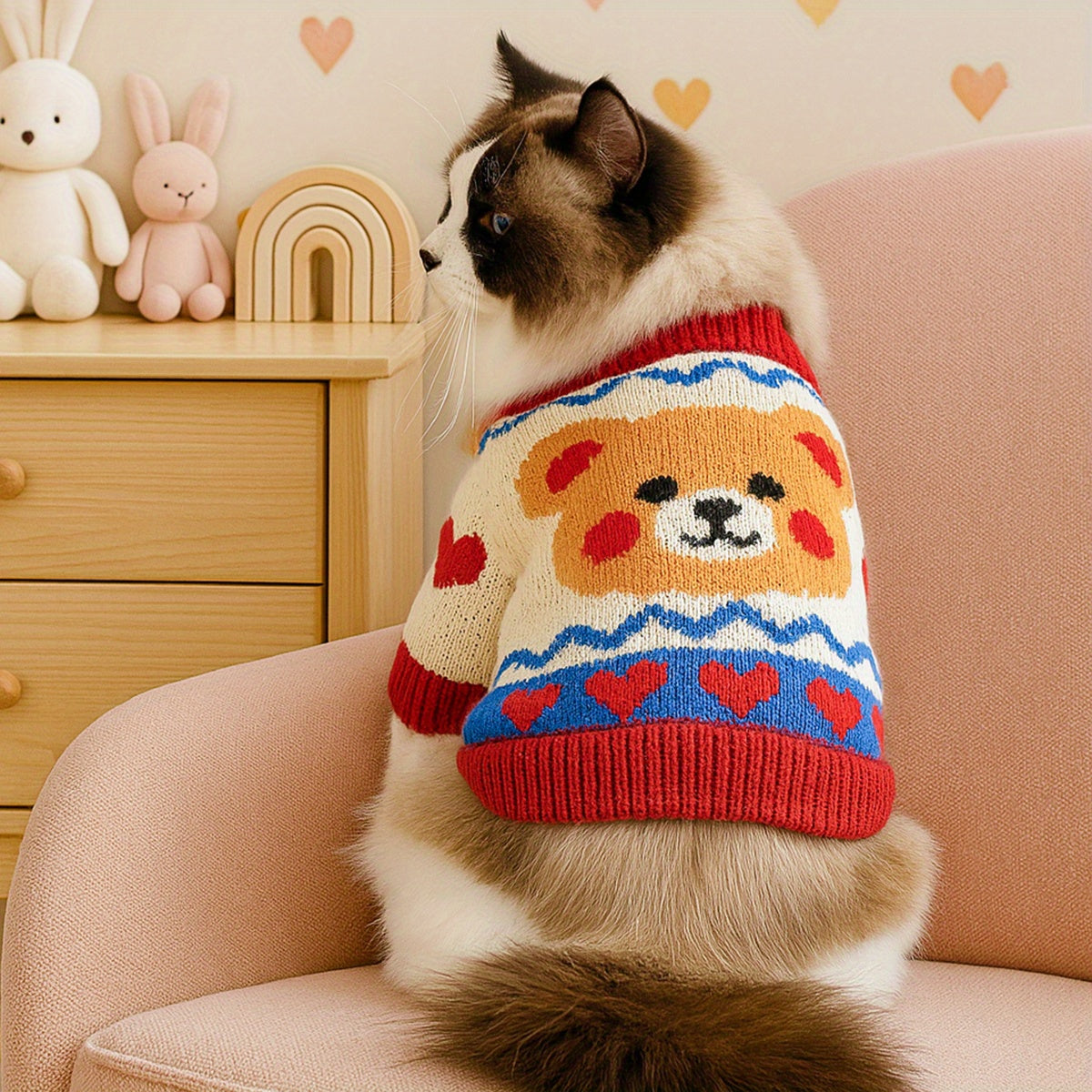 Cat wearing a colorful sweater with a bear design, sitting on a pink chair.