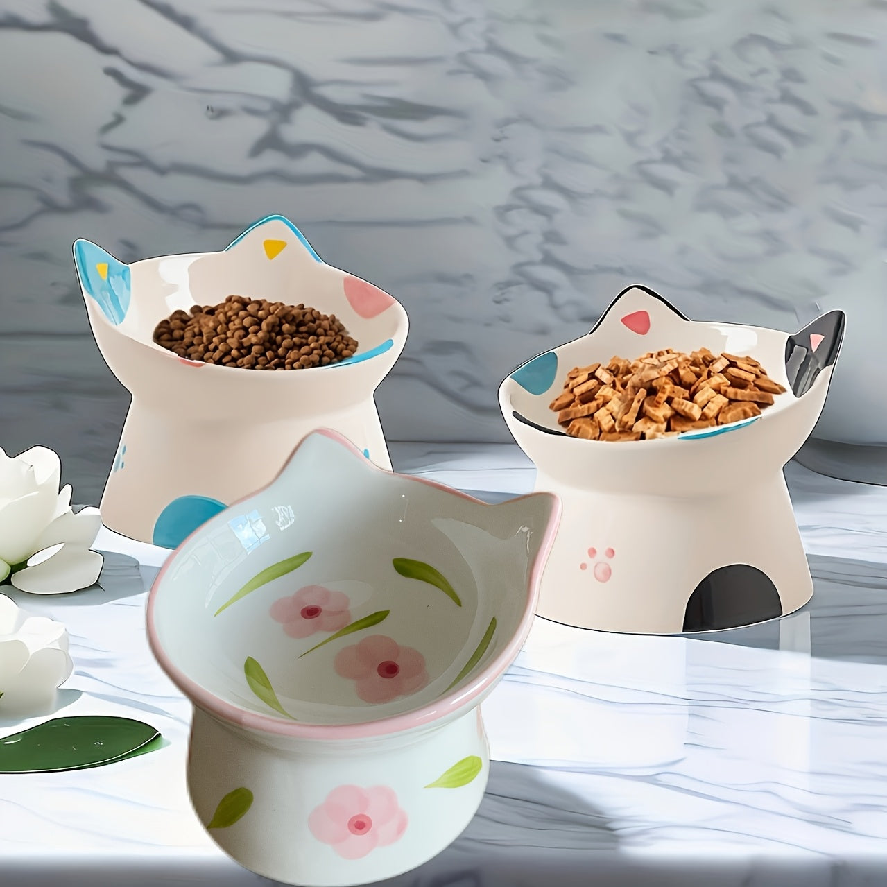 Three cat-shaped ceramic bowls with pet food on a marble surface.