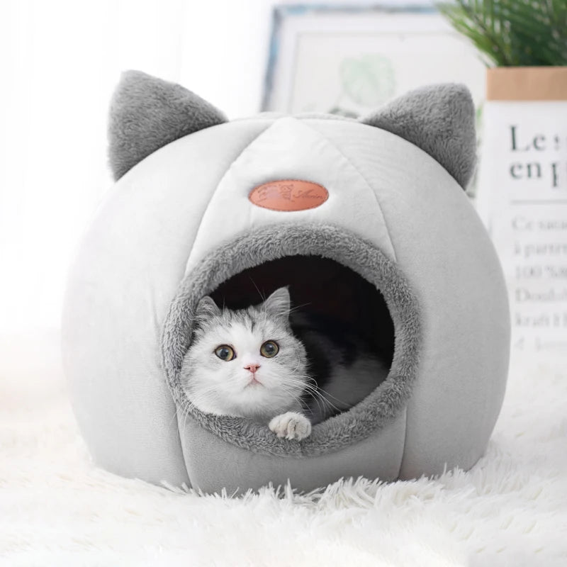 Cat peeking out from a gray and white pet bed with cat ears on a white surface.