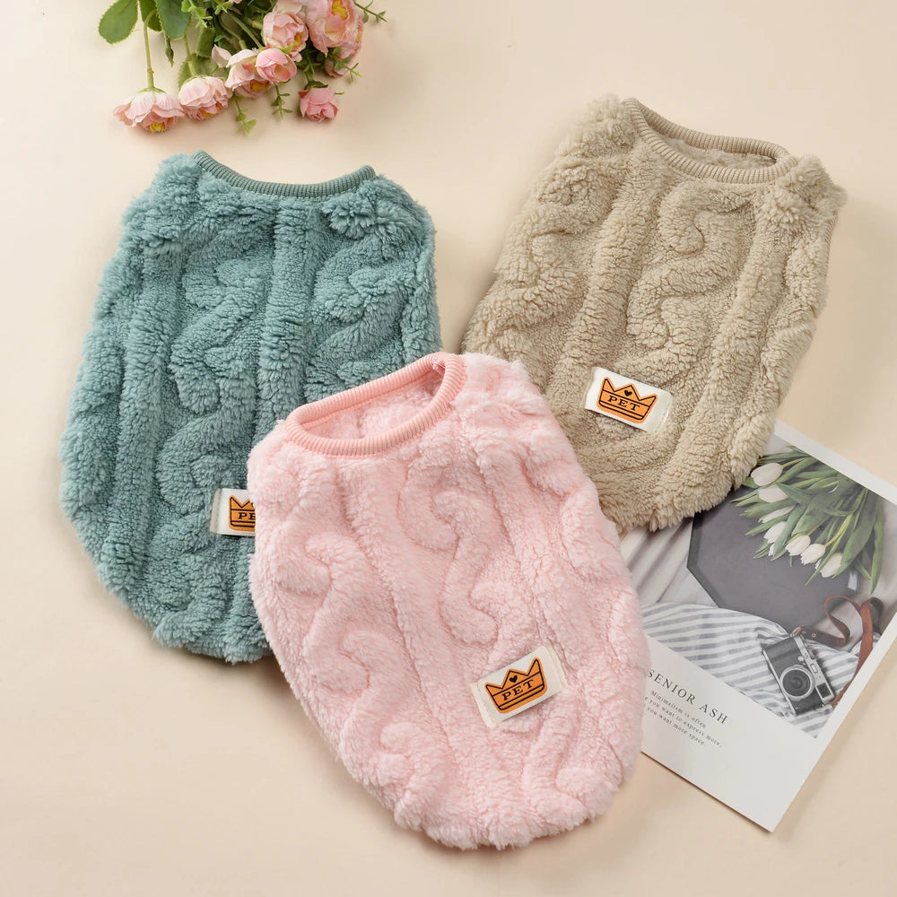 Three cable knit dog sweaters in green, pink, and beige on a light background with flowers and a magazine.