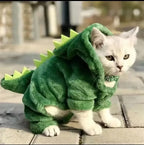 Cat wearing a green dinosaur costume on a wooden floor.
