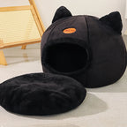Black cat-shaped pet bed on a light gray floor with a wooden chair in the background.