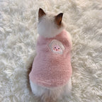 Small dog wearing a pink fleece sweater with a cartoon face on a fluffy white surface