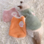 Three colorful pet vests with cartoon face designs on a fluffy white surface.