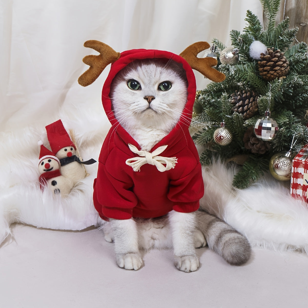 Cat wearing a red hoodie with reindeer antlers in front of a Christmas tree.