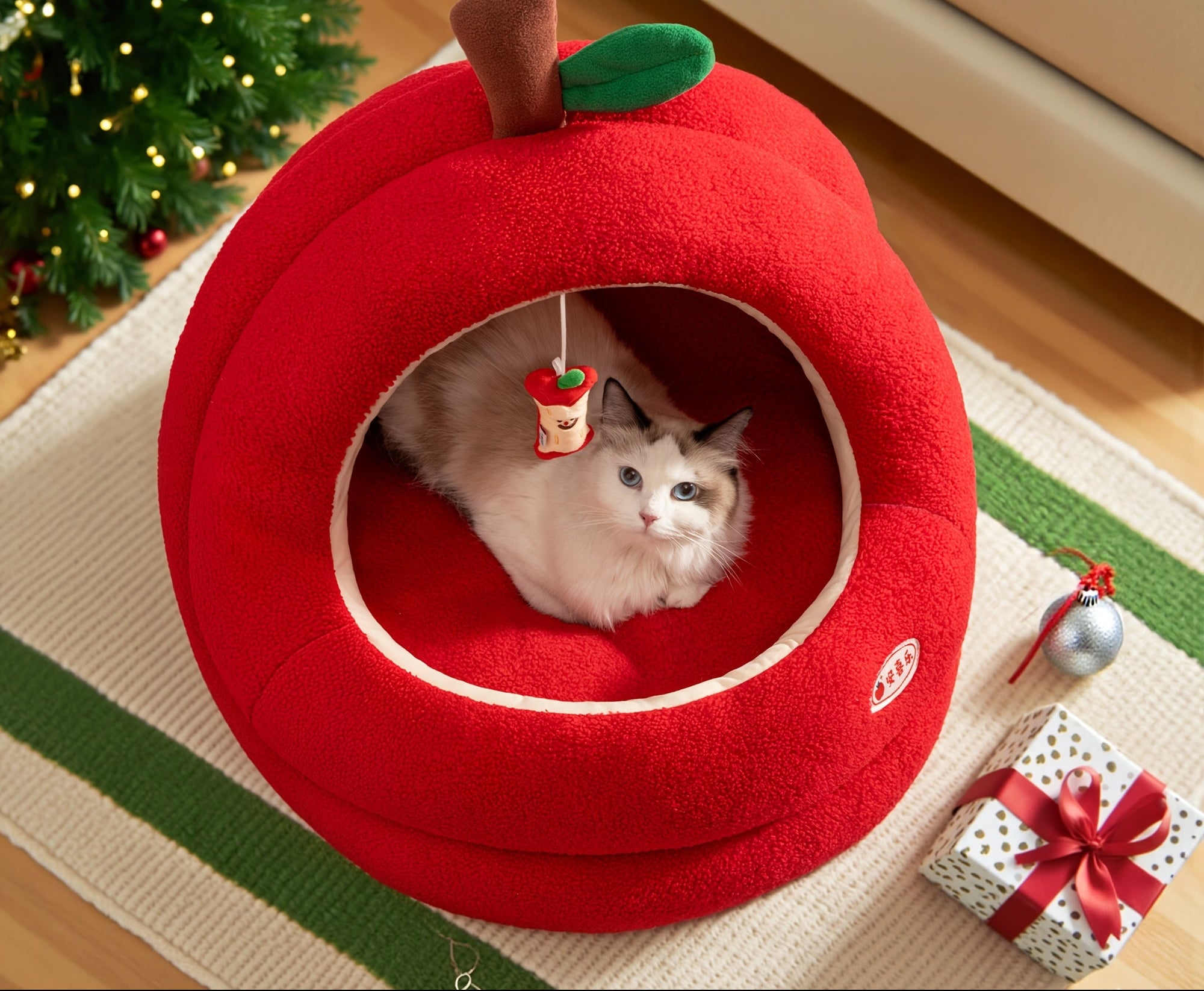 Red apple-shaped pet bed with a cat inside, Christmas tree and decorations in the background