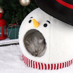 Cat sleeping in a snowman-shaped pet bed with Christmas decorations in the background.